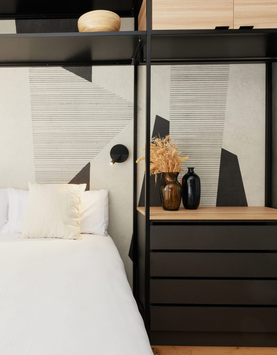 Bed, geometric headboard and black shelving with vases in a BelVillage Gavà apartment.