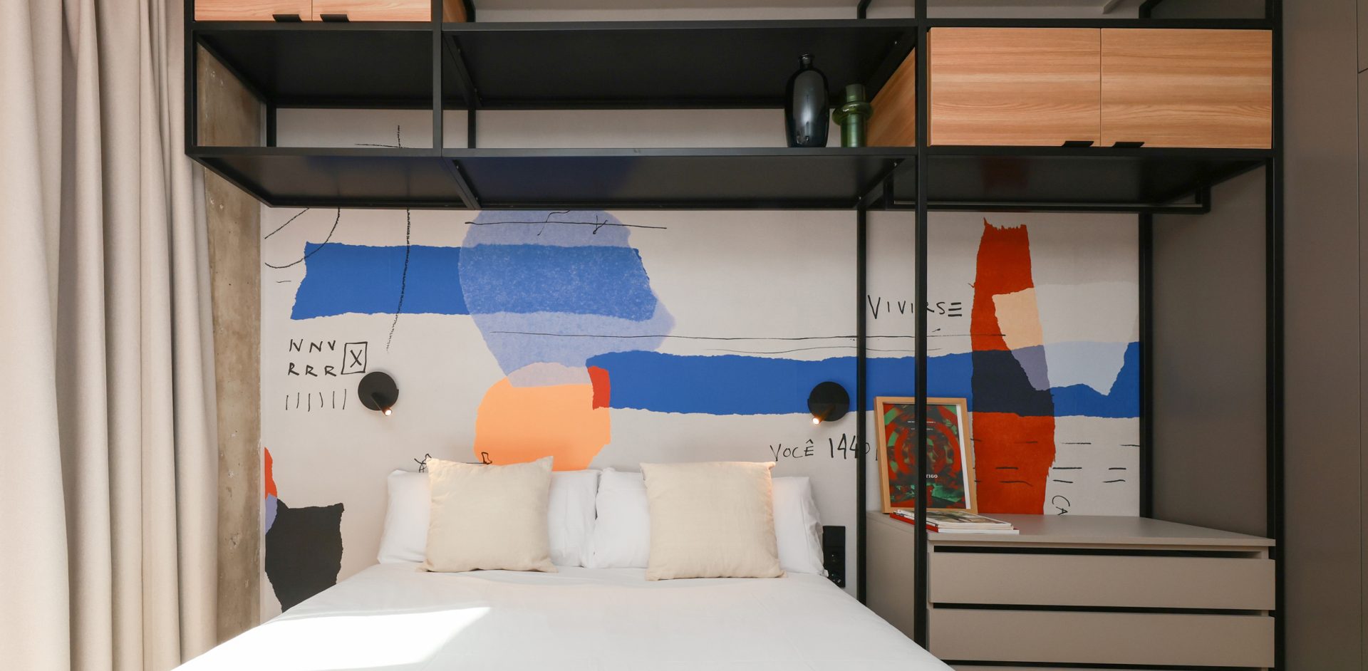Double bed beneath colourful artwork and black metal shelving in a Gavà apartment.